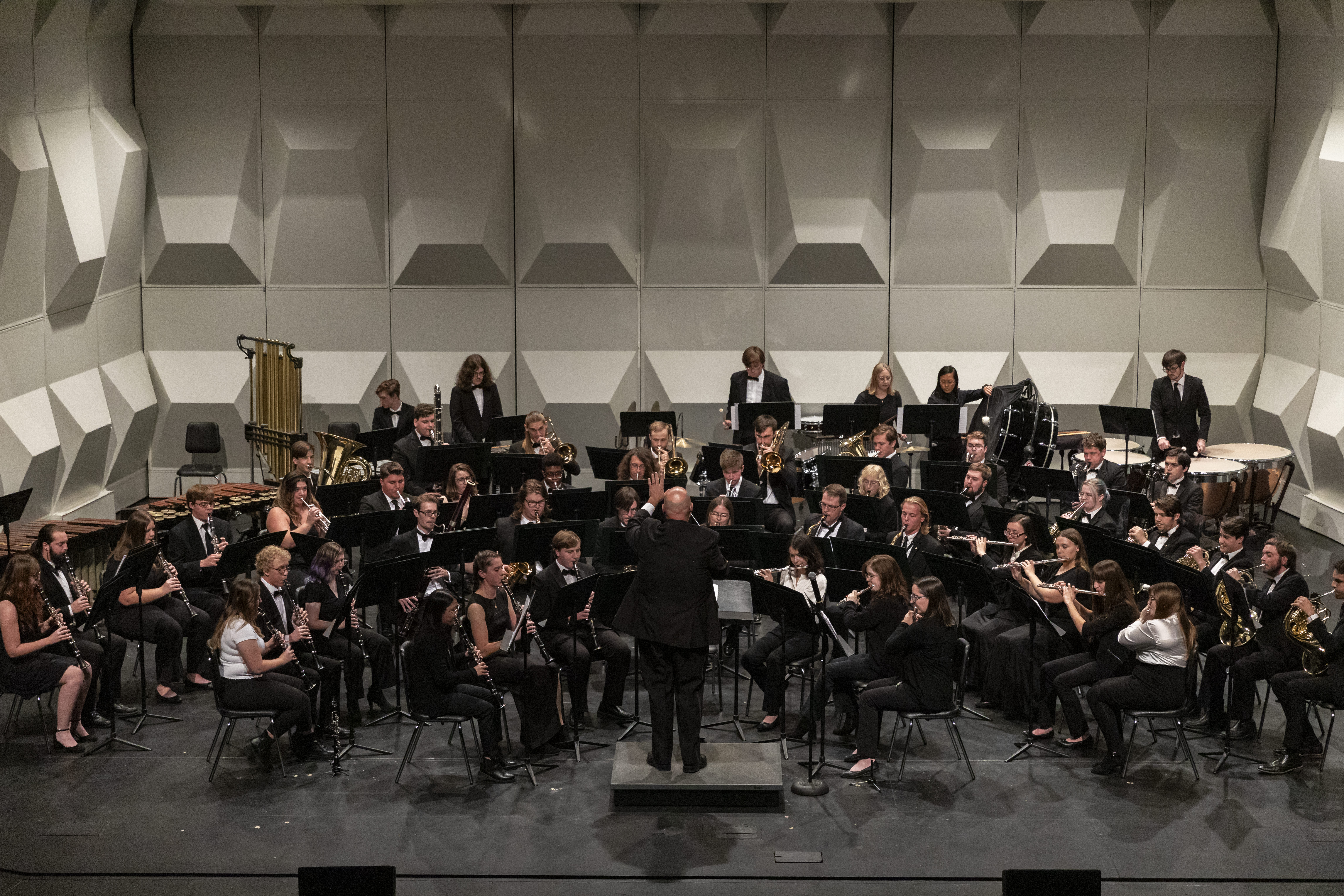 A concert band performs on stage with a conductor leading musicians. The ensemble, dressed in formal black attire, plays various brass and woodwind instruments.